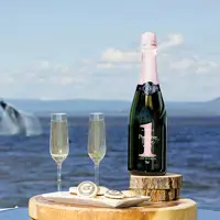 Une bouteille de vin mousseux et deux verres remplis reposent sur un plateau en bois près de l'océan. À proximité, des pâtisseries gourmandes sont disposées. En arrière-plan, une baleine jaillit de l'eau sous un ciel partiellement nuageux.