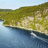 Vue aérienne d'un ferry naviguant à travers une rivière sinueuse bordée de forêts. Le navire laisse une traînée dans l'eau bleu profond, tandis que des falaises rocheuses et des arbres denses et verdoyants longent les rives sous un ciel clair.