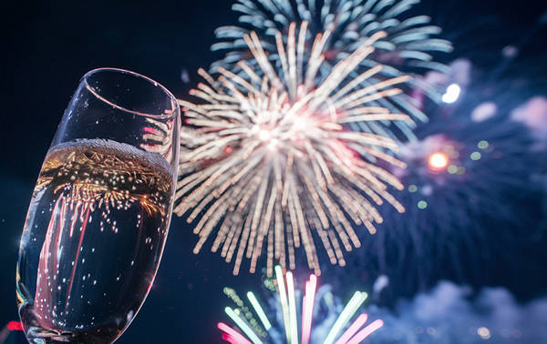 Un verre de champagne levé au premier plan tandis que des feux d'artifice multicolores illuminent le ciel nocturne en arrière-plan.