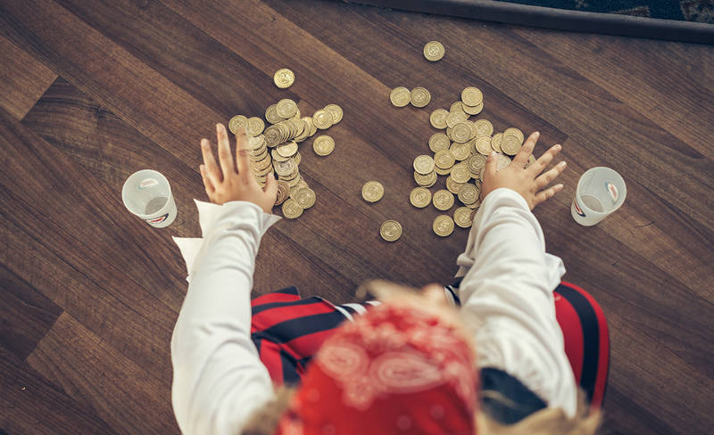 A person dressed as a pirate is sitting on a wooden floor, organizing gold coins into piles. Two white cups are nearby. The person is wearing a red bandana and pirate-themed clothing.