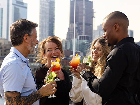 Un groupe de quatre amis souriants trinque avec des cocktails colorés sur une terrasse, avec des gratte-ciel en arrière-plan.