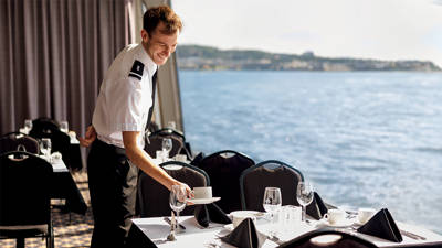 Un serveur en uniforme prépare une table élégante avec des nappes blanches et des serviettes noires dans une salle à manger avec une vue panoramique sur l'eau en arrière-plan.