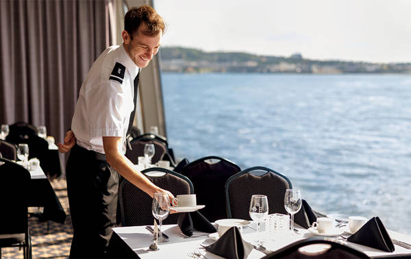 Un serveur en uniforme prépare une table élégante avec des nappes blanches et des serviettes noires dans une salle à manger avec une vue panoramique sur l'eau en arrière-plan.