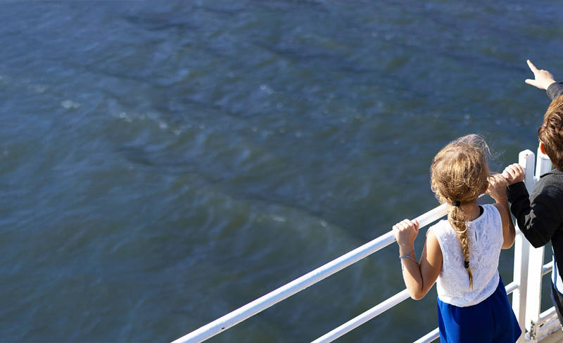 Two children stand on the pier of an AML cruise ship, looking out over the water. One child points at something in the distance. They are next to a white railing, with the sun shining brightly.