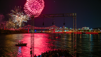 A vibrant fireworks display in a black sky above the AML Cavalier Maxim ship in Montreal, with the city illuminated in the distance.