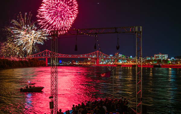 Un feu d'artifice vibrant dans un ciel noir au-dessus du navire AML Cavalier Maxim à Montréal, avec la ville illuminée au loin.