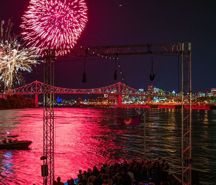 Un feu d'artifice vibrant dans un ciel noir au-dessus du navire AML Cavalier Maxim à Montréal, avec la ville illuminée au loin.