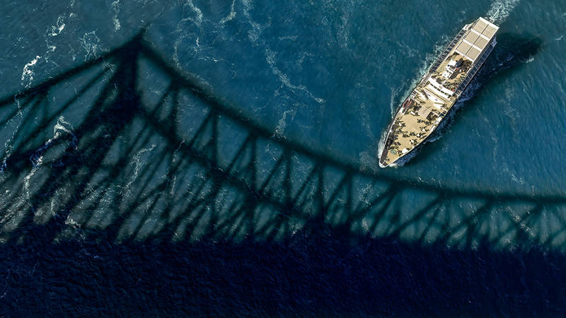 Vue aérienne d'un bateau naviguant sous un pont, dont l'ombre se projette sur la surface bleue de l'eau.