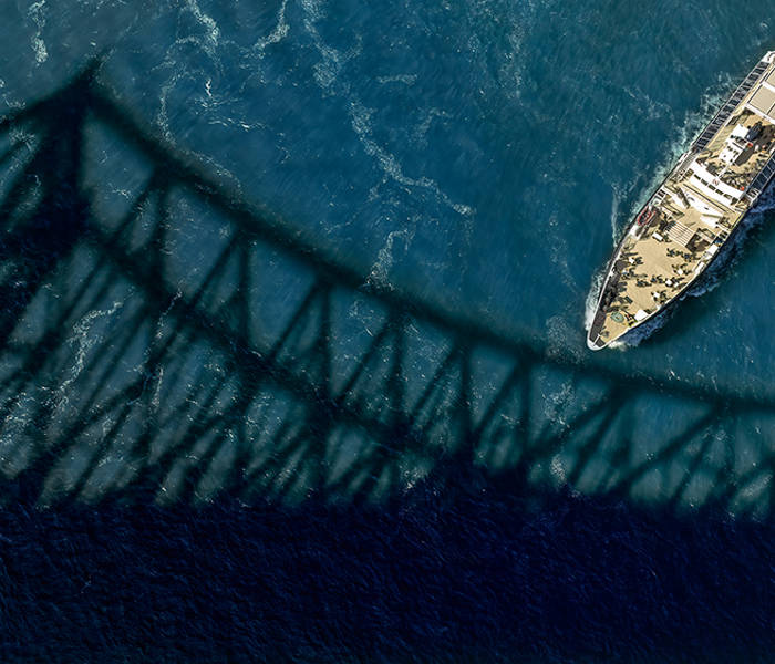 Vue aérienne d'un bateau naviguant sous un pont, dont l'ombre se projette sur la surface bleue de l'eau.