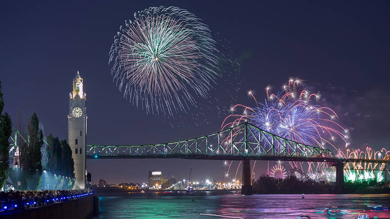 Une vue nocturne spectaculaire d'un feu d'artifice illuminant le ciel au-dessus d'un pont emblématique et d'une tour horloge, avec des lumières urbaines scintillantes et une rivière calme au premier plan.