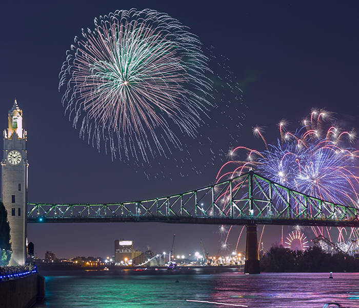 Une vue nocturne spectaculaire d'un feu d'artifice illuminant le ciel au-dessus d'un pont emblématique et d'une tour horloge, avec des lumières urbaines scintillantes et une rivière calme au premier plan.