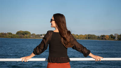 Une femme de dos, portant une blouse noire et des lunettes de soleil, appuyée contre une rambarde, admirant une rivière avec des arbres et un ciel dégagé en arrière-plan.