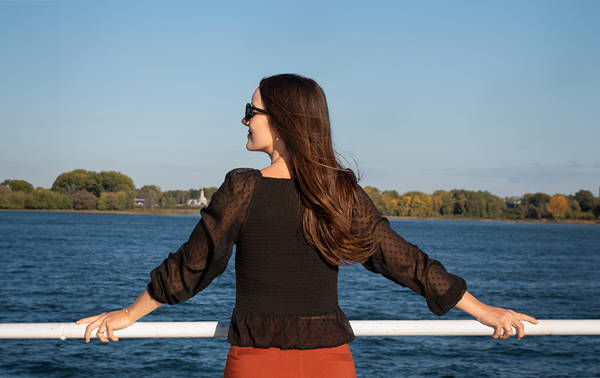 Une femme de dos, portant une blouse noire et des lunettes de soleil, appuyée contre une rambarde, admirant une rivière avec des arbres et un ciel dégagé en arrière-plan.