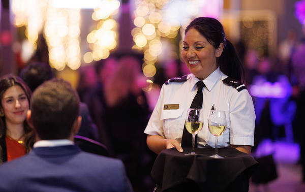  Une serveuse en uniforme souriante sert deux verres de vin blanc à une table dans un environnement élégant et animé, illuminé par des lumières décoratives floues en arrière-plan.