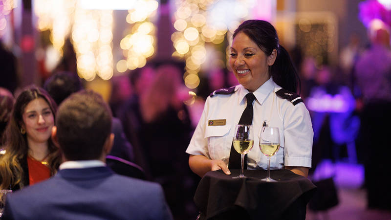  Une serveuse en uniforme souriante sert deux verres de vin blanc à une table dans un environnement élégant et animé, illuminé par des lumières décoratives floues en arrière-plan.