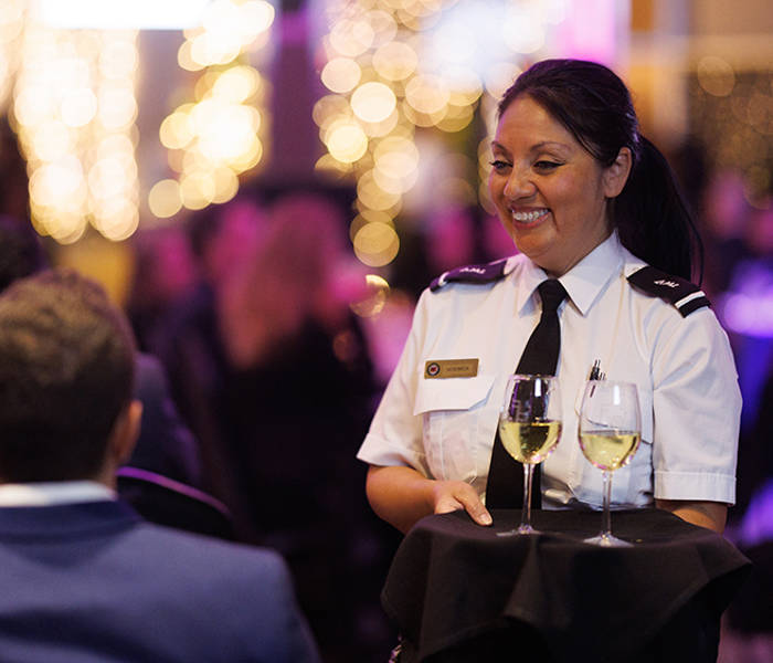  Une serveuse en uniforme souriante sert deux verres de vin blanc à une table dans un environnement élégant et animé, illuminé par des lumières décoratives floues en arrière-plan.