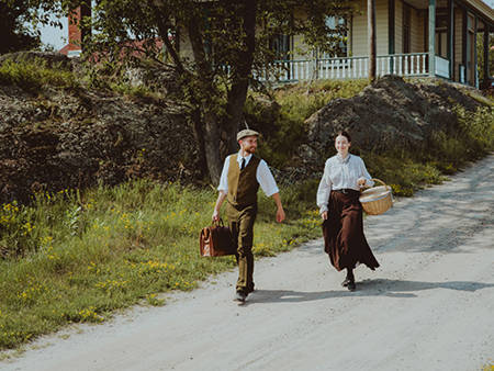 Two characters dressed in period costume are walking on Grosse Île. They are descending a small slope, with a historic house in the background.