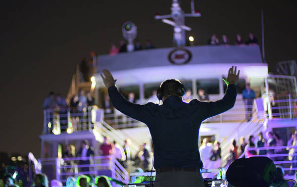 Un DJ anime une fête nocturne sur un bateau, avec des lumières colorées et une foule dansante sur plusieurs niveaux du navire.