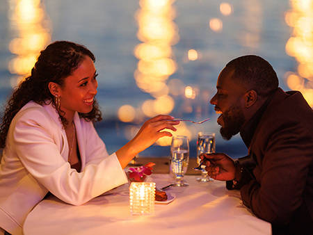 Un couple partage un moment romantique lors d'un dîner aux chandelles près de l'eau. La femme, souriante, donne une bouchée de dessert à l'homme, avec des lumières floues scintillant en arrière-plan.