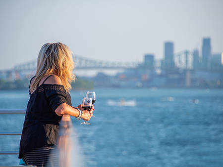 Une femme blonde, de dos, tenant un verre de vin rouge, regardant une rivière avec un pont et des gratte-ciel en arrière-plan par une journée ensoleillée.