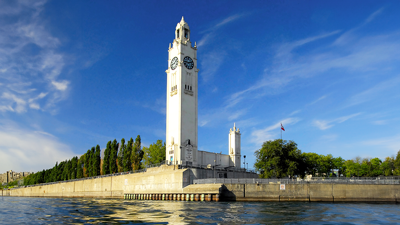 View of the Clock Tower in the Old Port of Montreal from the St. Lawrence River, under a blue sky with a few clouds.