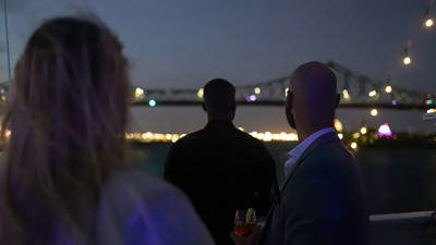 Trois personnes vues de dos, profitant d'une soirée sur un bateau avec un pont illuminé et les lumières de la ville en arrière-plan.