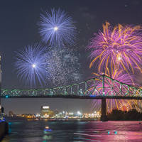 Un feu d'artifice coloré éclate dans le ciel nocturne près du pont Jacques-Cartier et de la tour de l'Horloge, avec des reflets sur le fleuve et une foule observant depuis le quai.