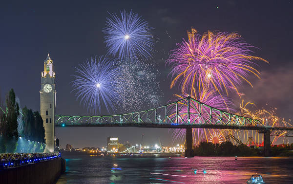 Un feu d'artifice coloré éclate dans le ciel nocturne près du pont Jacques-Cartier et de la tour de l'Horloge, avec des reflets sur le fleuve et une foule observant depuis le quai.