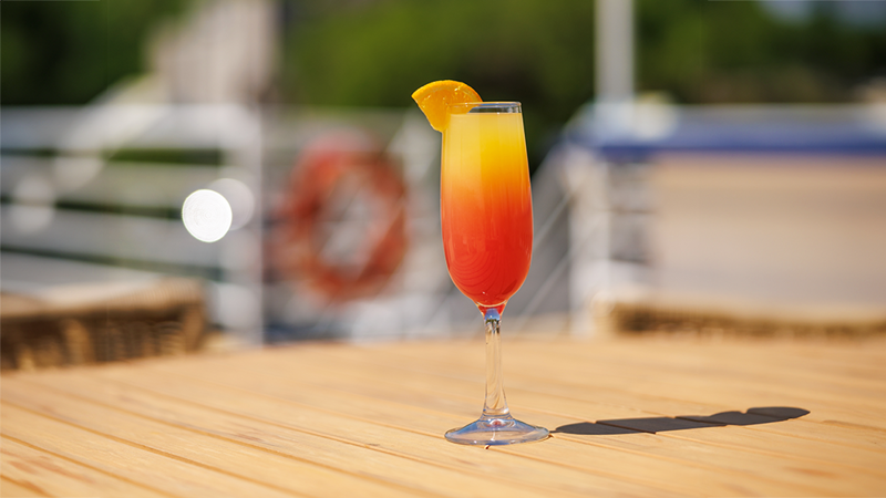 Un cocktail mimosa orangé dans une flûte avec un quartier d'orange sur le dessus du verre, servi sur une table en bois située sur l'une des terrasses extérieures du navire AML Cavalier Maxim à Montréal. 