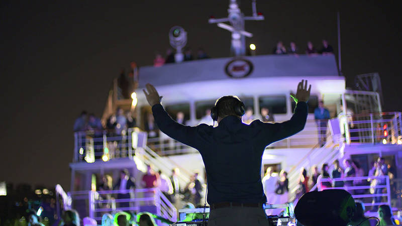 Un DJ anime une fête nocturne sur un bateau, avec des lumières colorées et une foule dansante sur plusieurs niveaux du navire.