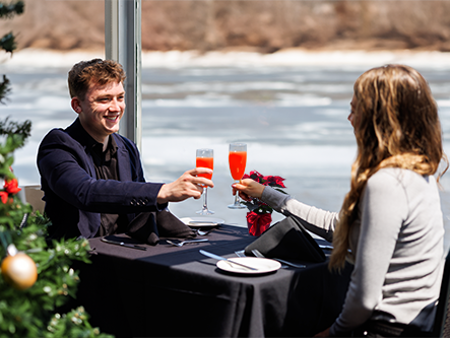 Couple complice et souriant en train de trinquer à table avec un cocktail lors d'une croisière brunch de Noël avec un sapin de Noël décoré au premier plan à gauche