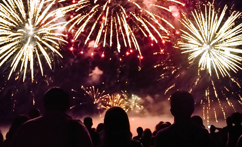 A crowd of spectators watches a spectacular fireworks display lighting up the night sky with golden and red bursts. The scene is filled with wonder and celebration.