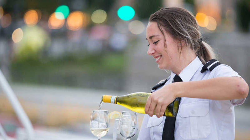 Une serveuse souriante en uniforme verse du vin blanc dans des verres, avec un arrière-plan flou de lumières et de verdure.