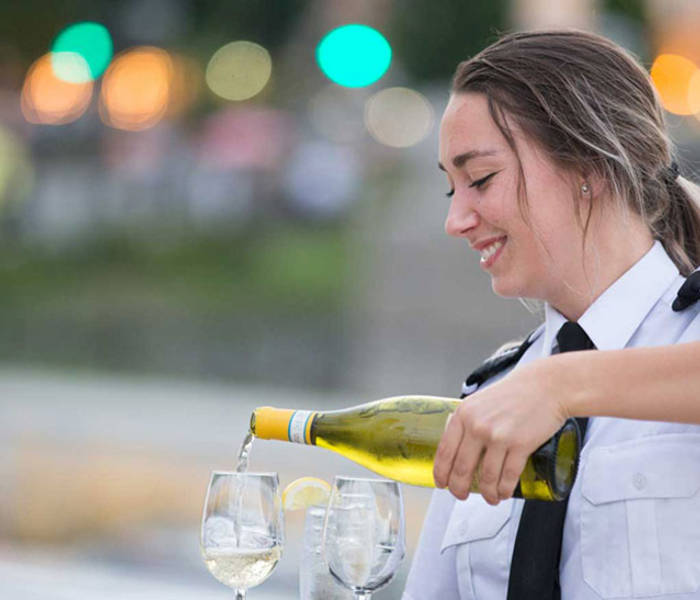 Une serveuse souriante en uniforme verse du vin blanc dans des verres, avec un arrière-plan flou de lumières et de verdure.