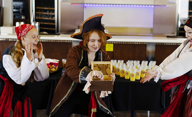 Three people dressed as pirates are playfully interacting indoors. One holds a wooden chest with a surprised expression, while the other two react with excitement. They are near a table with multiple bottles, suggesting a playful gathering or event.