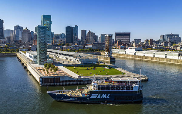 Vue aérienne d'un bateau de croisière naviguant près d'un quai moderne avec des espaces verts et des sculptures, entouré de bâtiments urbains et d'une rivière sous un ciel dégagé.