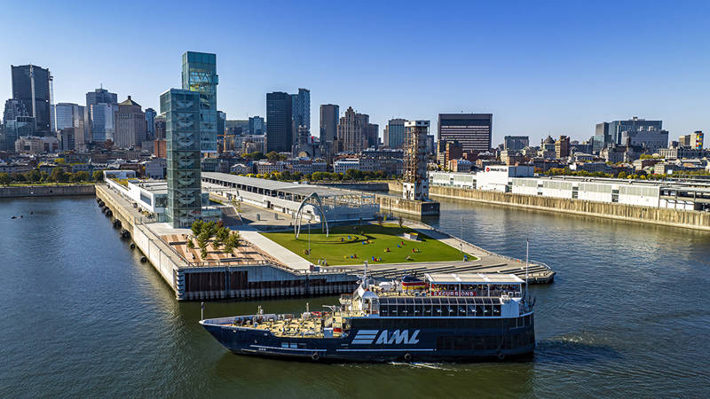 Vue aérienne d'un bateau de croisière naviguant près d'un quai moderne avec des espaces verts et des sculptures, entouré de bâtiments urbains et d'une rivière sous un ciel dégagé.