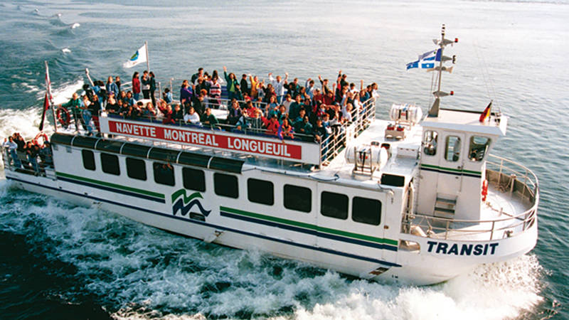 Une navette fluviale pleine de passagers profite d'une traversée entre Montréal et Longueuil sur des eaux calmes, avec des drapeaux flottants sur le bateau.