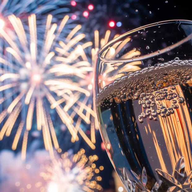 A sparkling glass of champagne is in the foreground, capturing the reflections of a spectacular fireworks display lighting up the night sky in the background.