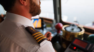 Gros plan sur un capitaine en uniforme avec des galons dorés sur l'épaule, dans la cabine de pilotage d'un bateau, entouré d'équipements de navigation et d'instruments.