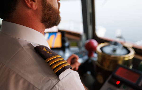 Gros plan sur un capitaine en uniforme avec des galons dorés sur l'épaule, dans la cabine de pilotage d'un bateau, entouré d'équipements de navigation et d'instruments.