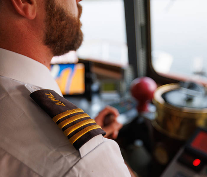 Gros plan sur un capitaine en uniforme avec des galons dorés sur l'épaule, dans la cabine de pilotage d'un bateau, entouré d'équipements de navigation et d'instruments.