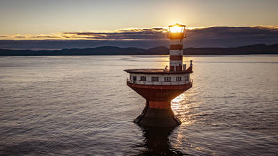 Un phare solitaire, rayé de bandes, se dresse dans des eaux calmes au coucher du soleil. Le soleil brille intensément derrière la lanterne, diffusant une lueur chaleureuse. À l’arrière-plan, des collines sombres et lointaines se découpent sous un ciel partiellement nuageux, ajoutant une touche dramatique à la scène.