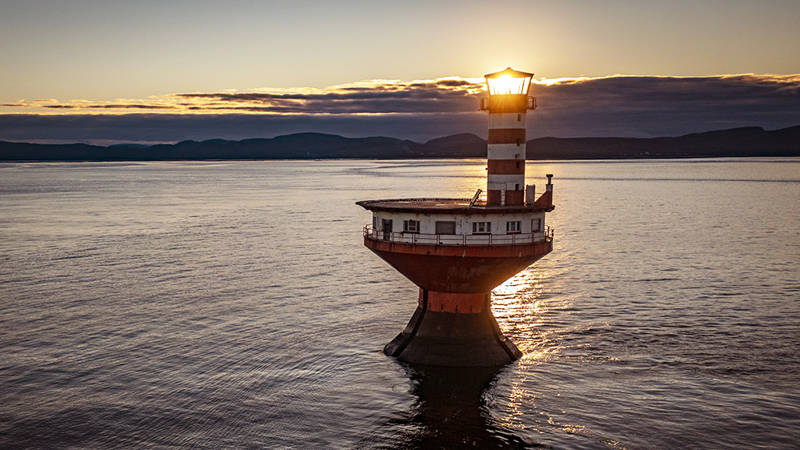 Un phare solitaire, rayé de bandes, se dresse dans des eaux calmes au coucher du soleil. Le soleil brille intensément derrière la lanterne, diffusant une lueur chaleureuse. À l’arrière-plan, des collines sombres et lointaines se découpent sous un ciel partiellement nuageux, ajoutant une touche dramatique à la scène.