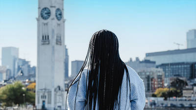 Une femme de dos avec de longues tresses noires, regardant une grande tour horloge blanche et une ville avec des bâtiments modernes sous un ciel bleu dégagé.