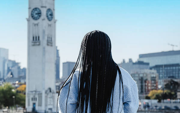 Une femme de dos avec de longues tresses noires, regardant une grande tour horloge blanche et une ville avec des bâtiments modernes sous un ciel bleu dégagé.