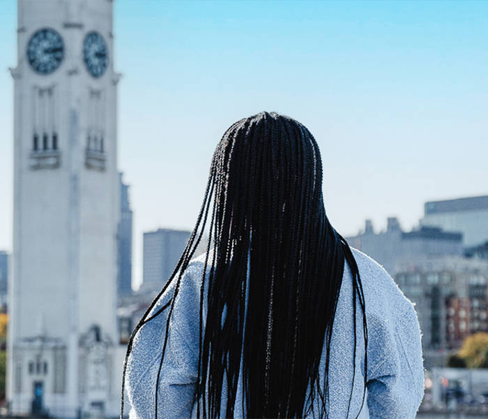 Une femme de dos avec de longues tresses noires, regardant une grande tour horloge blanche et une ville avec des bâtiments modernes sous un ciel bleu dégagé.