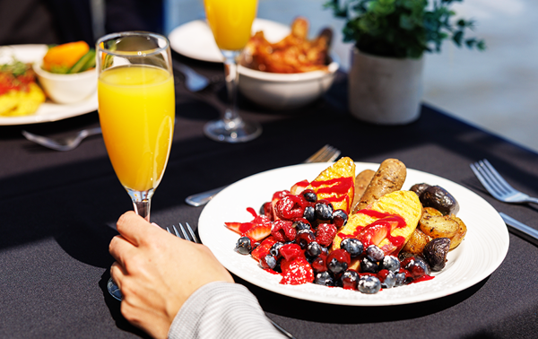  A close-up of a vegan brunch plate with vegan sausage, fruits, and baby potatoes, with a hand holding a mimosa glass.