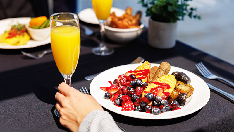 Gros plan sur une assiette brunch végane avec une saucisse végane, des fruits et des pommes de terre grelots avec une main tenant un verre de mimosa.