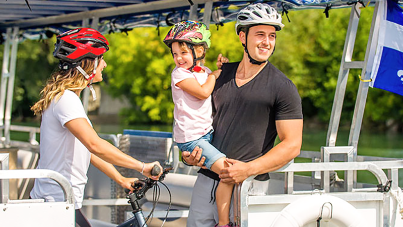 Un couple et un enfant profite d'une balade en navette fluviale sur le Saint-Laurent à Montréal. La femme tient un vélo et porte un casque sur la tête,  l'homme porte l'enfant dans ses bras.